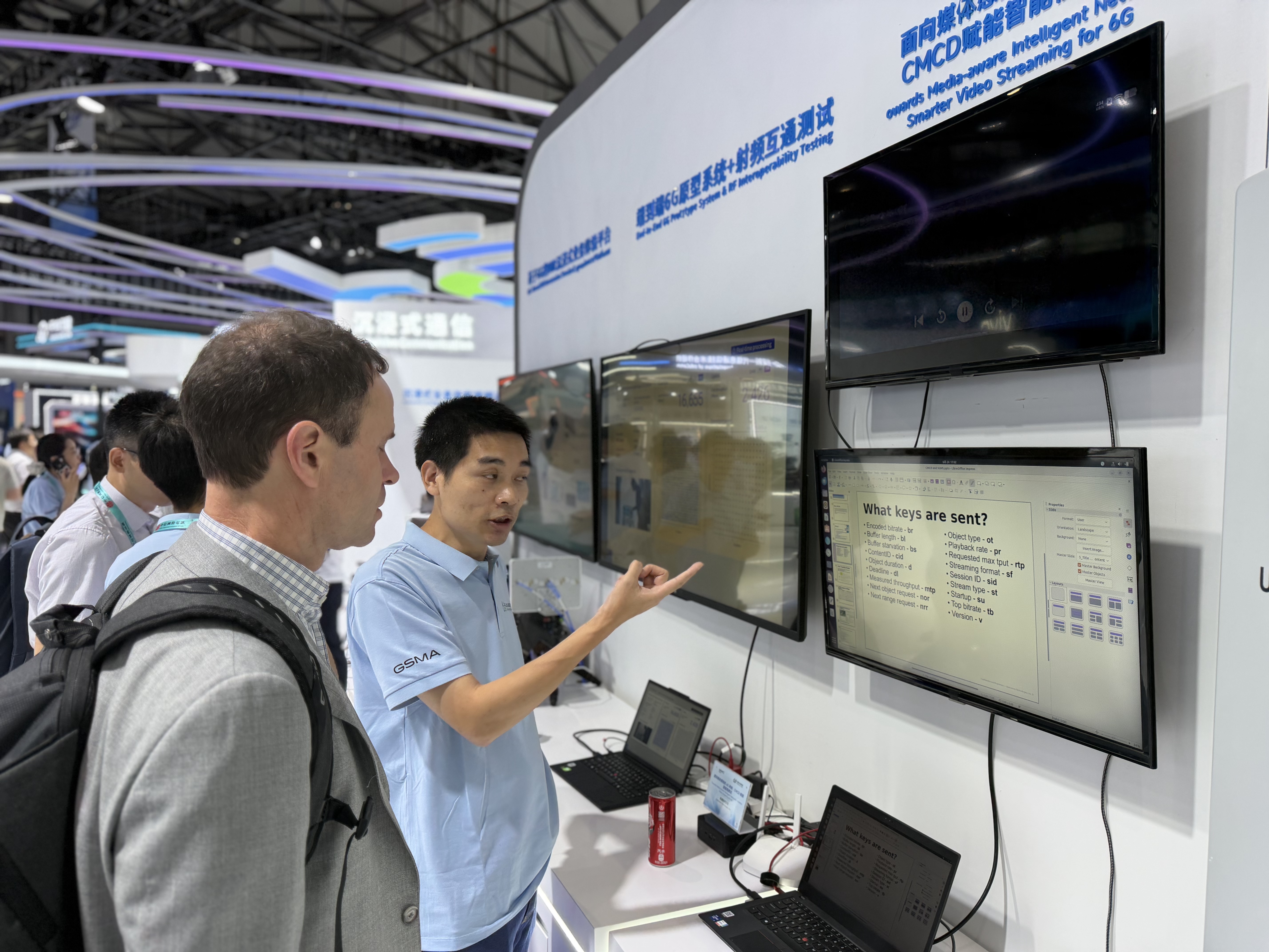 Demo presenter pointing to a screen showing CMCD key parameters, explaining the live streaming demo to a visitor at Qualcomm's booth, MWC Shanghai